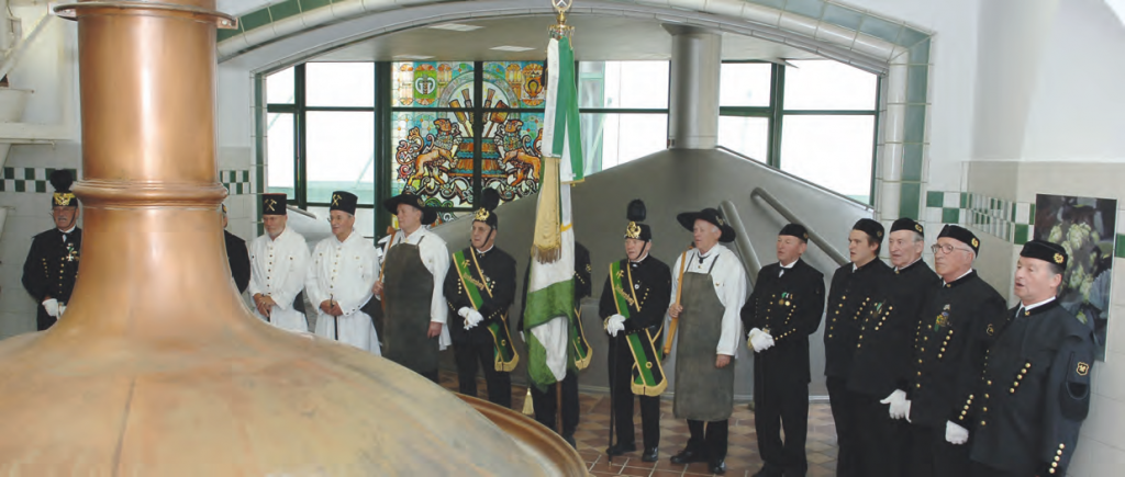 A group of men dressed in traditional brewer and mining uniforms stand in a semi-circle inside a historic brewery, singing in front of a large copper brew kettle. A stained glass window with brewery symbols is visible in the background. The event appears ceremonial and honors brewing traditions.