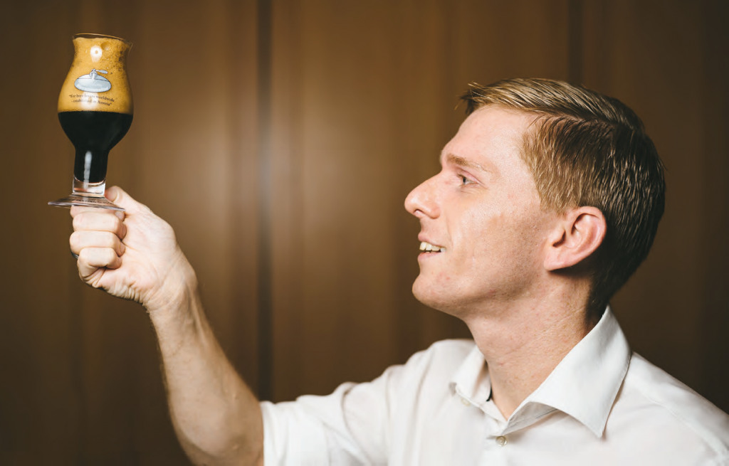 A man in a white shirt holds a tulip-shaped glass of dark beer up to the light, examining its color and foam.