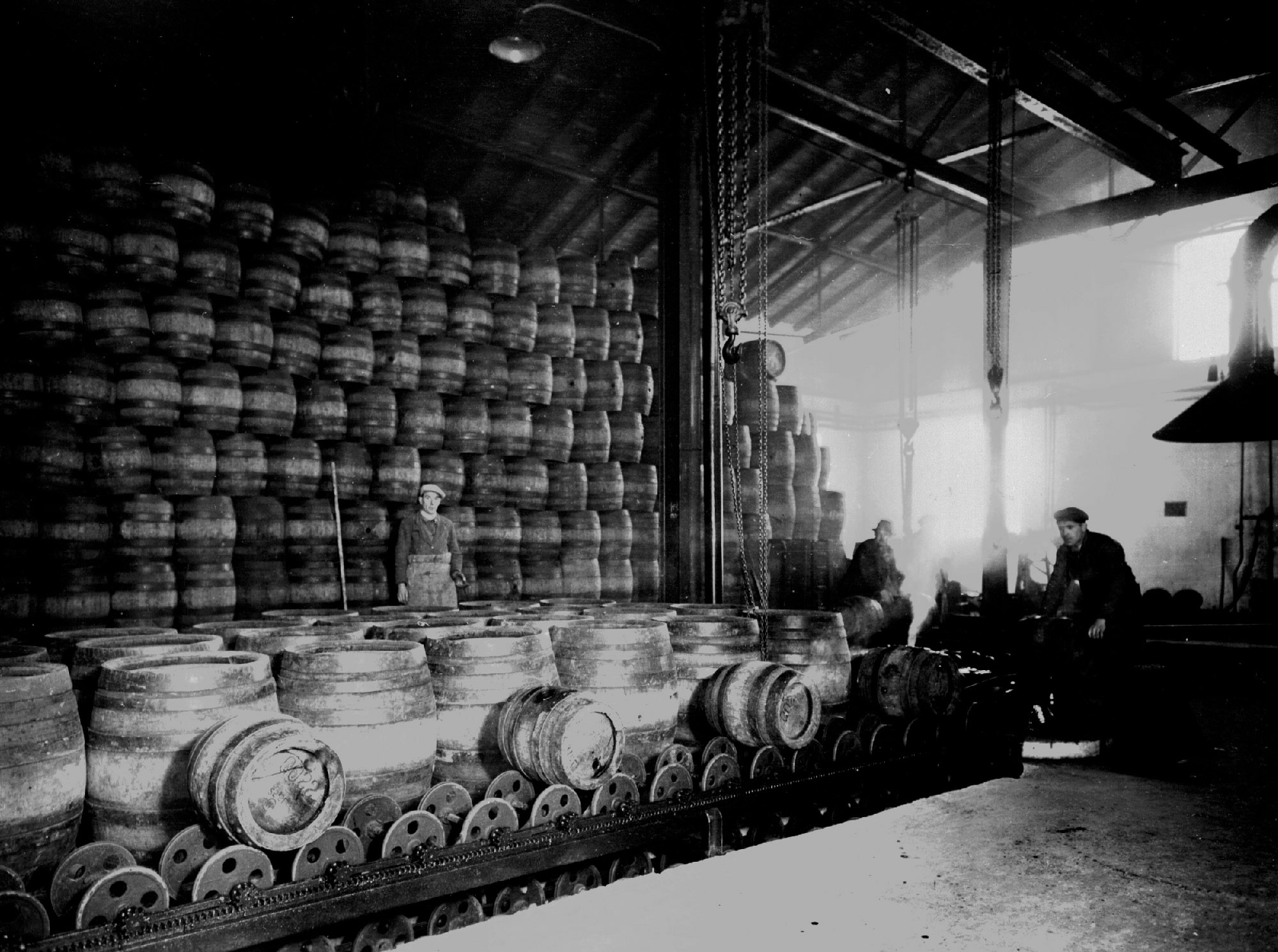 Workers in a historic brewery hall with a towering wall of wooden beer barrels and a conveyor system in the foreground.
