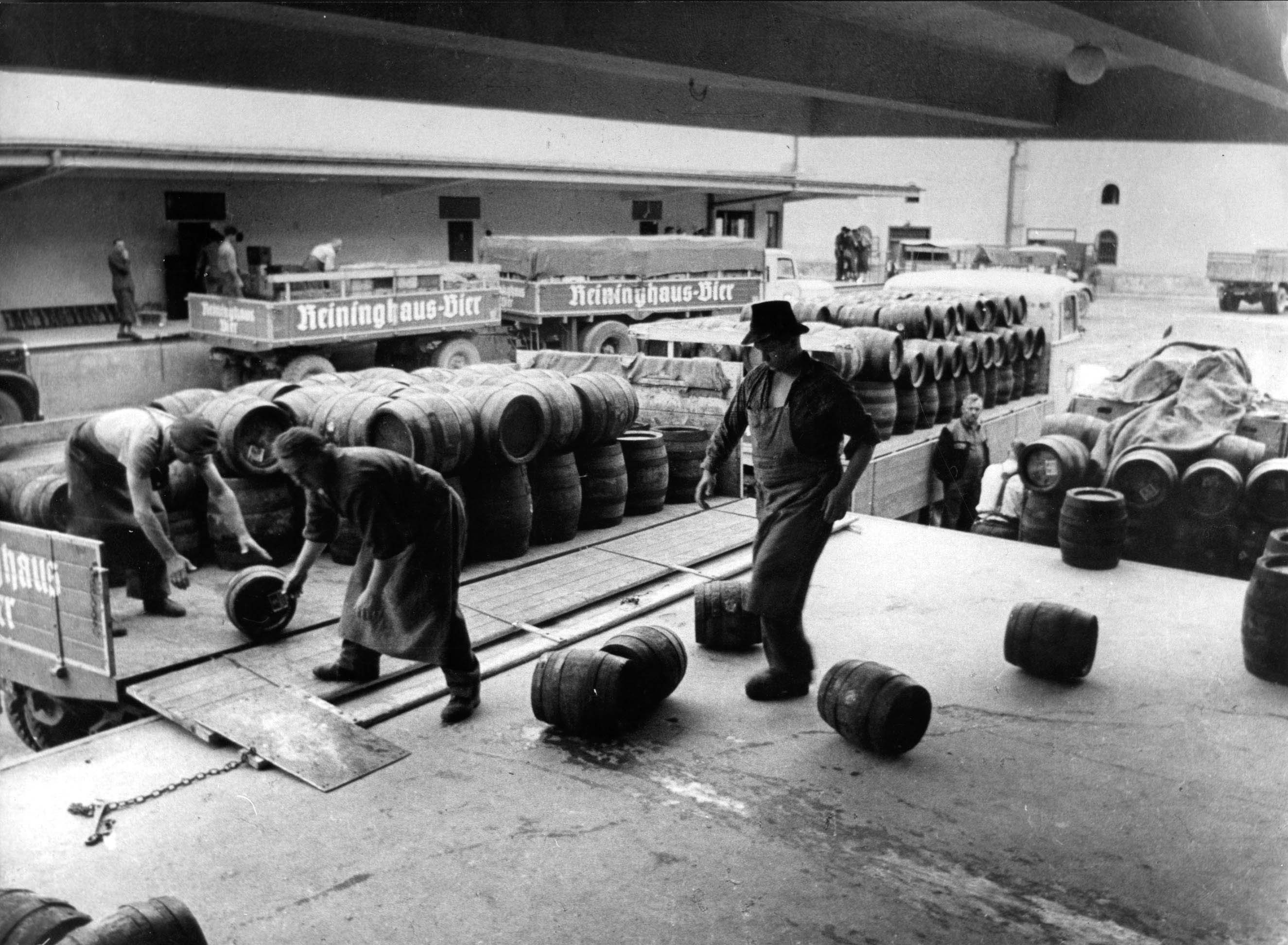 Workers loading wooden beer barrels onto trucks labeled ‘Reininghaus-Bier’ at a brewery.