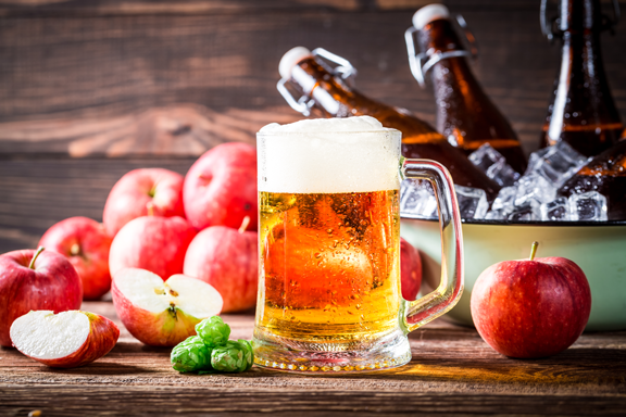 Refreshing glass of golden apple cider beer with a frothy head, surrounded by red apples, green hops, and swing-top bottles in an ice-filled bowl on a rustic wooden table.